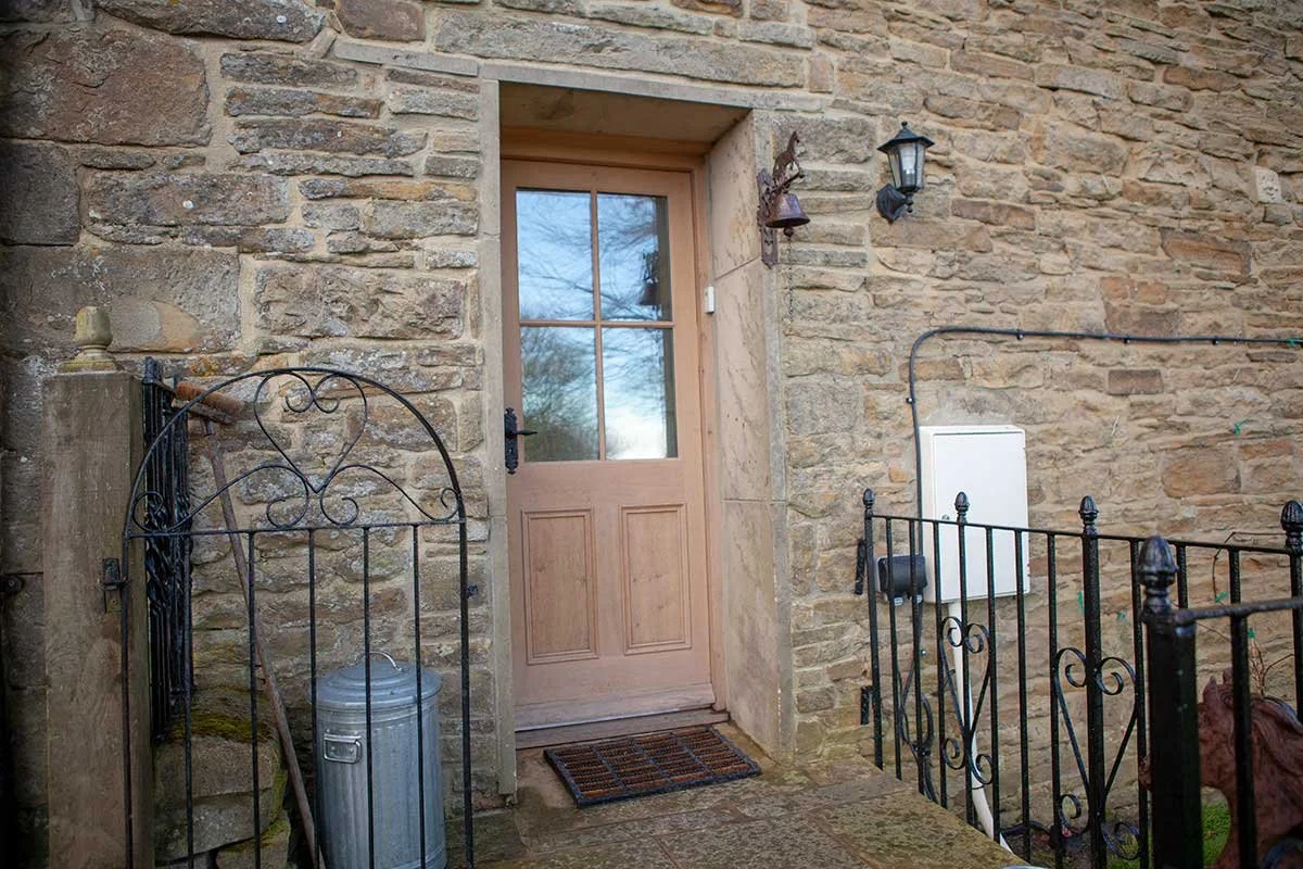 Wooden front door with window panels, surrounded by a stone wall, glass lantern and bell above, black metal railing and trash can nearby.