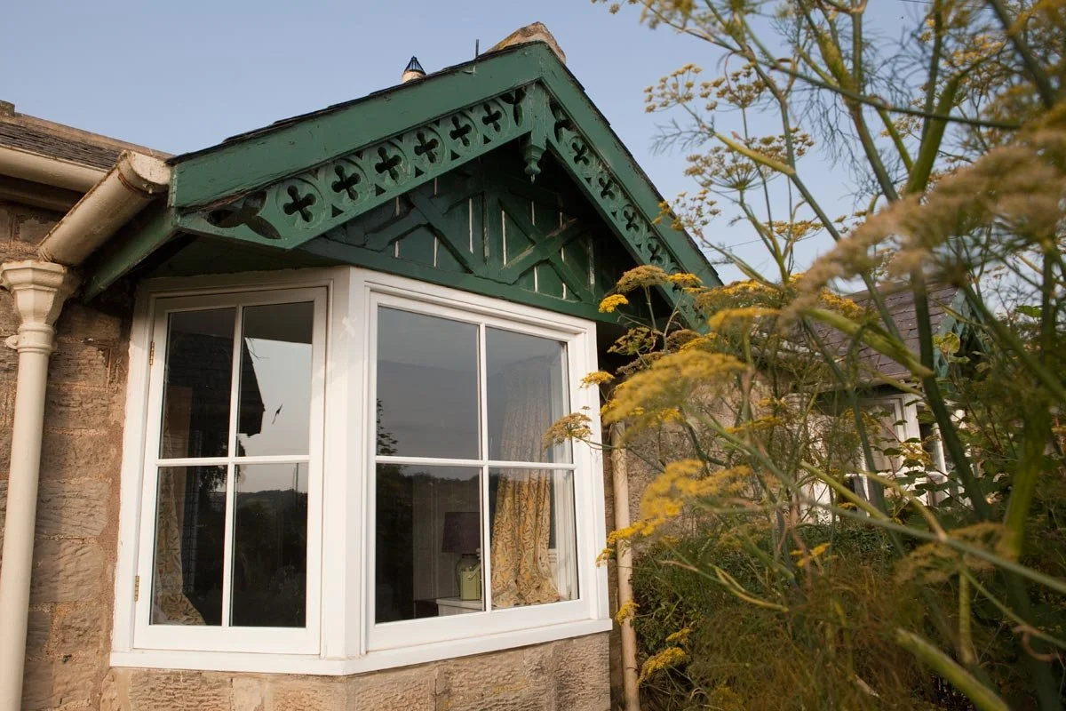A house exterior with a large cross-window and green decorative woodwork under the gable roof, surrounded by plants with yellow flowers.