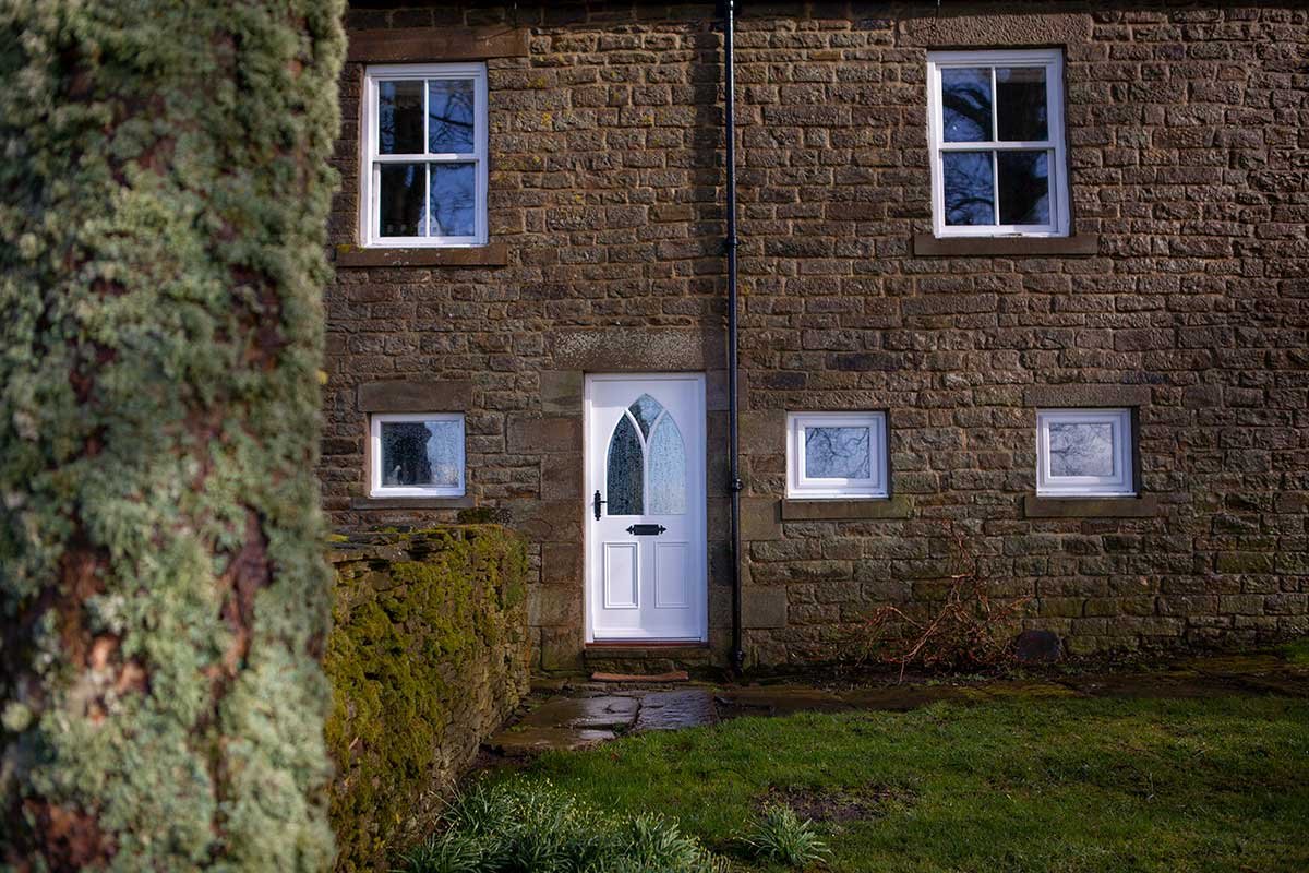 A brick house exterior with four windows and a white door with glass panels, viewed through a bush on the left side, with a small lawn and moss-covered wall in the foreground.