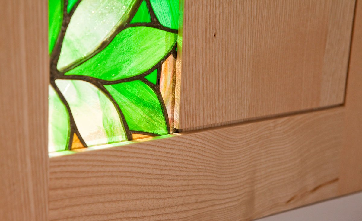 Close-up of a stained glass window with green and beige glass pieces, framed by light-colored wood.