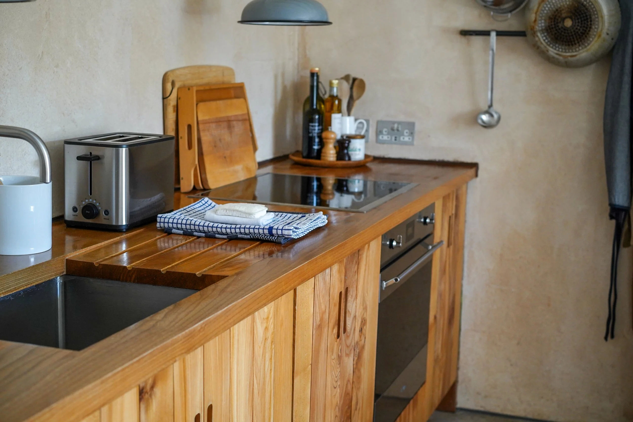 A rustic kitchen with wooden cabinets, a stove, and a countertop with a toaster, cutting boards, a towel, and a tray with bottles and cups.