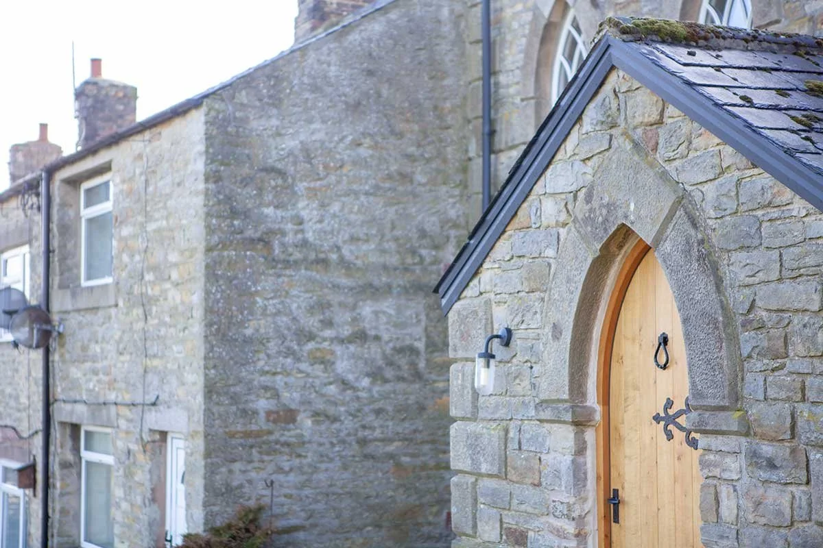 Stone building with an arched wooden door, Gothic-style metal hinges, black door handle, and a small light fixture outside. The roof has moss and is covered with dark shingles, and there are stone chimneys and windows visible on neighboring structure
