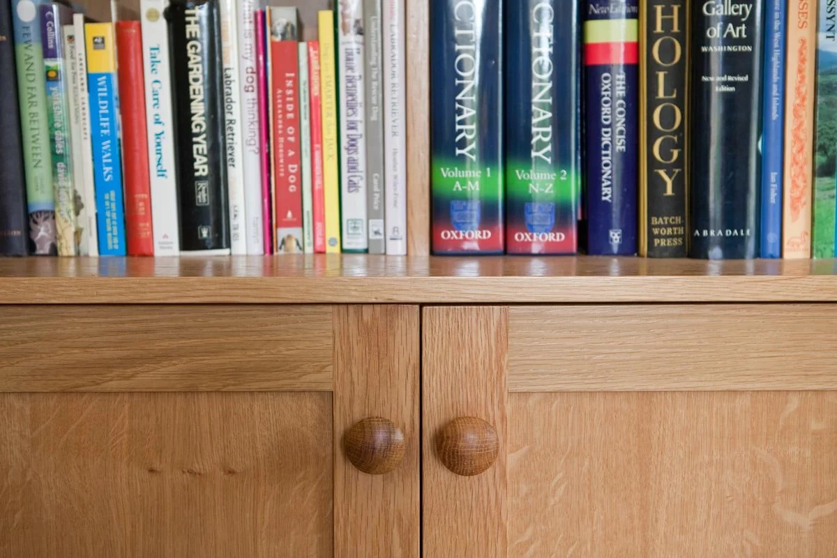 A wooden bookshelf with two closed cabinet doors at the bottom, featuring round wooden knobs. The top shelf is filled with a variety of books, including encyclopedias and art books, with some titles visible.