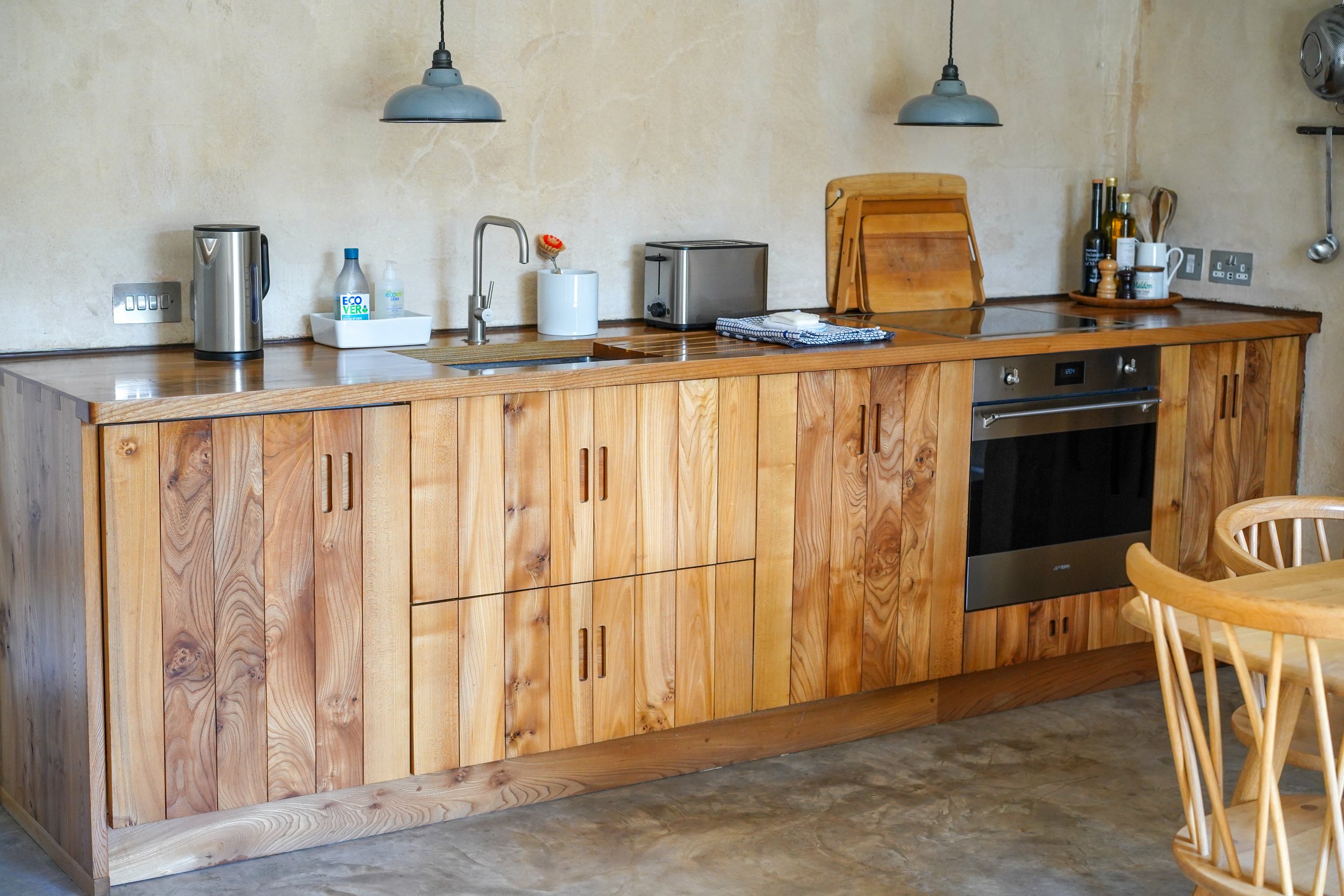 A kitchen with wooden cabinets, a stainless steel oven, a countertop with various kitchen items, pendant lights, and a kitchen chair.