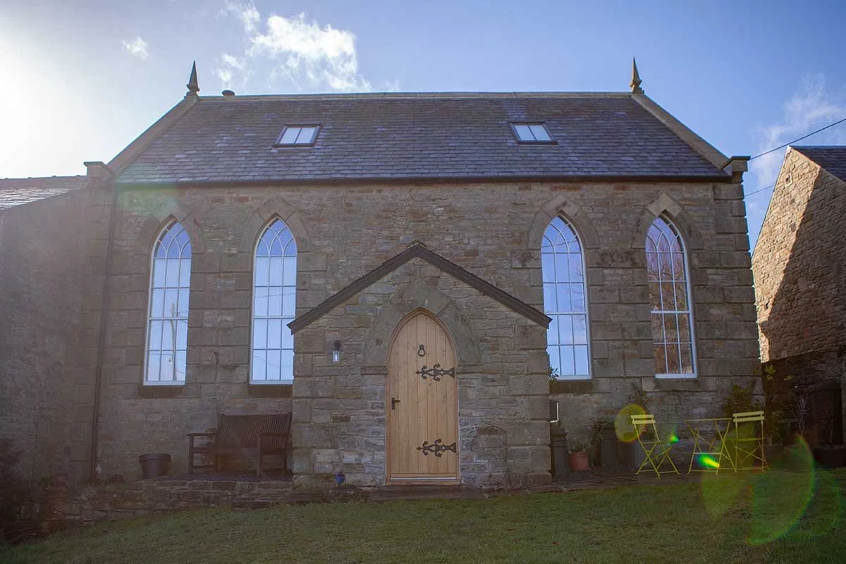 Front view of a stone church with large stained glass windows, a wooden door, and a small gabled porch in a grassy yard, with the sky and some clouds in the background.