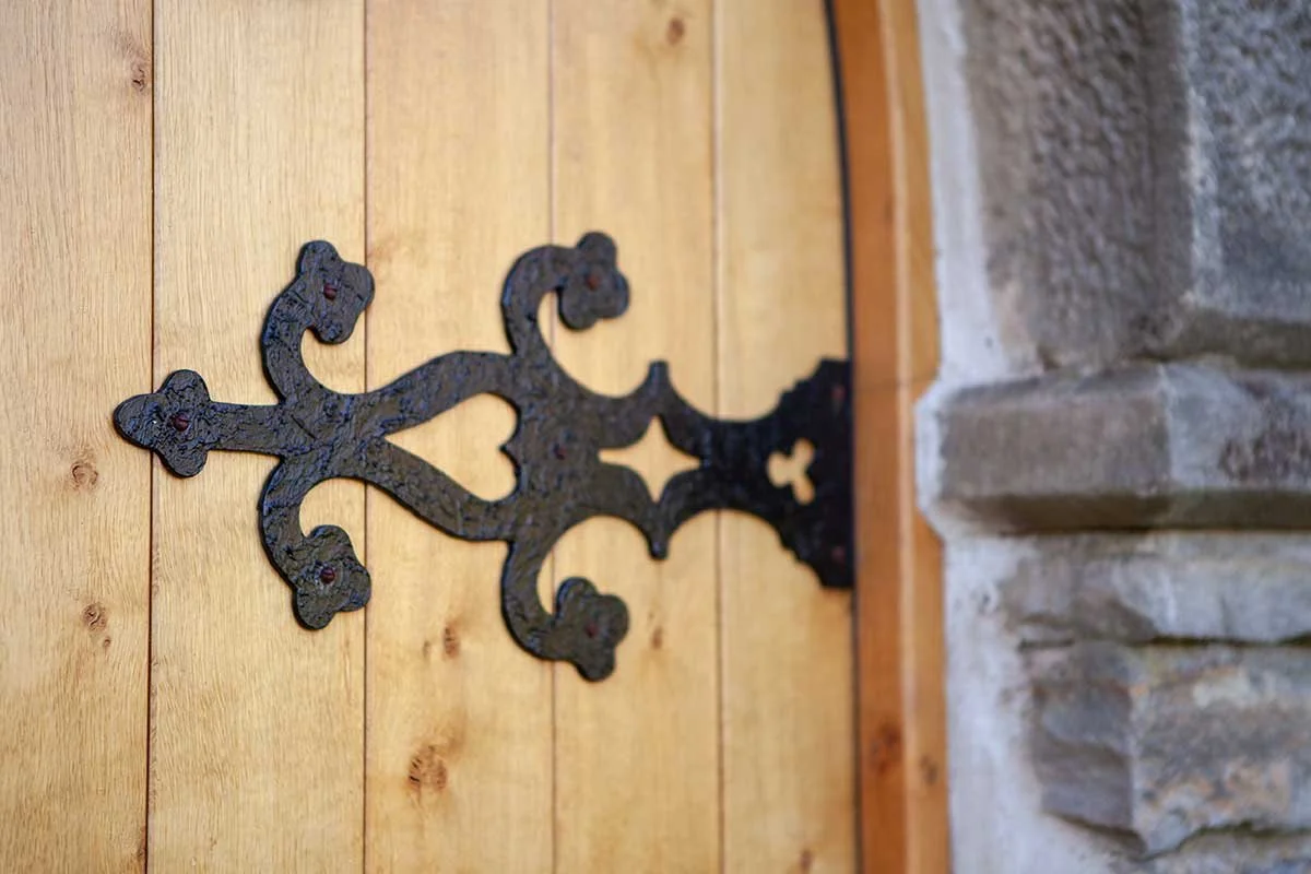 A close-up of decorative black wrought iron hinge attached to a wooden door, with a stone wall on the right side.