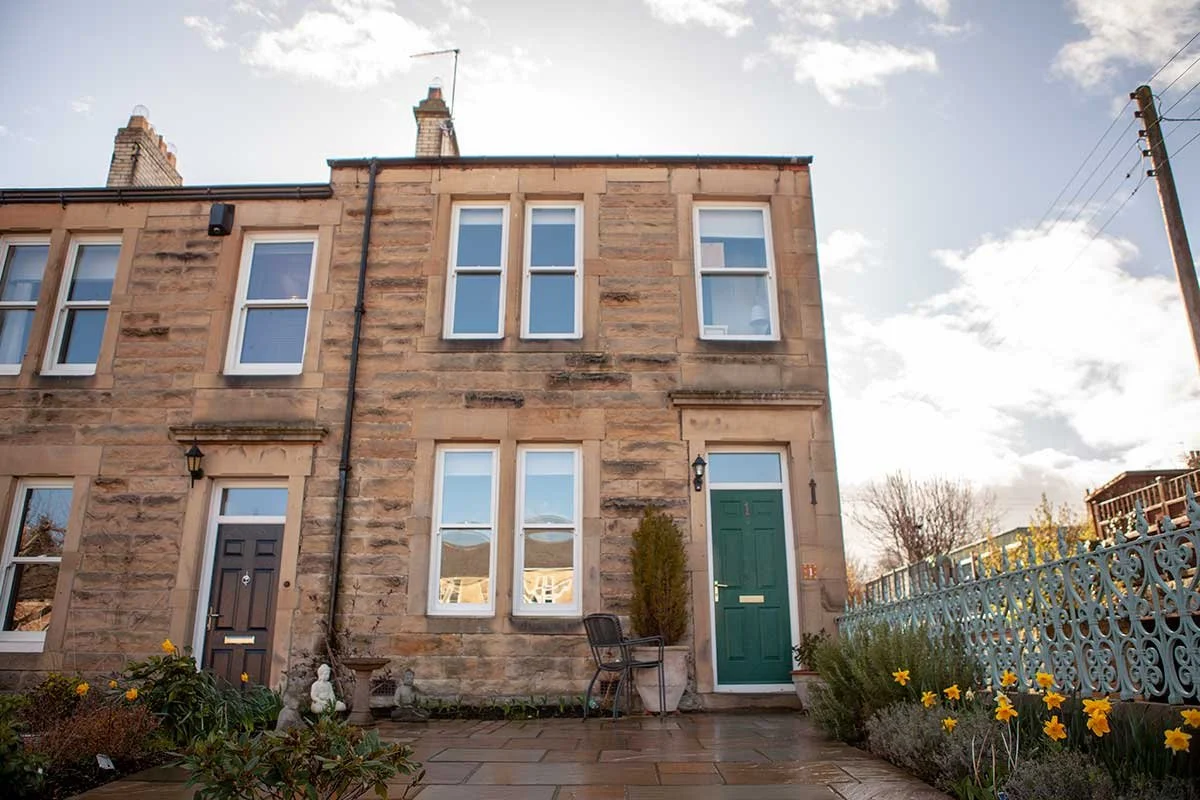 Front view of a sandstone house with a green door and multiple windows, surrounded by a garden with yellow flowers and a blue decorative fence.