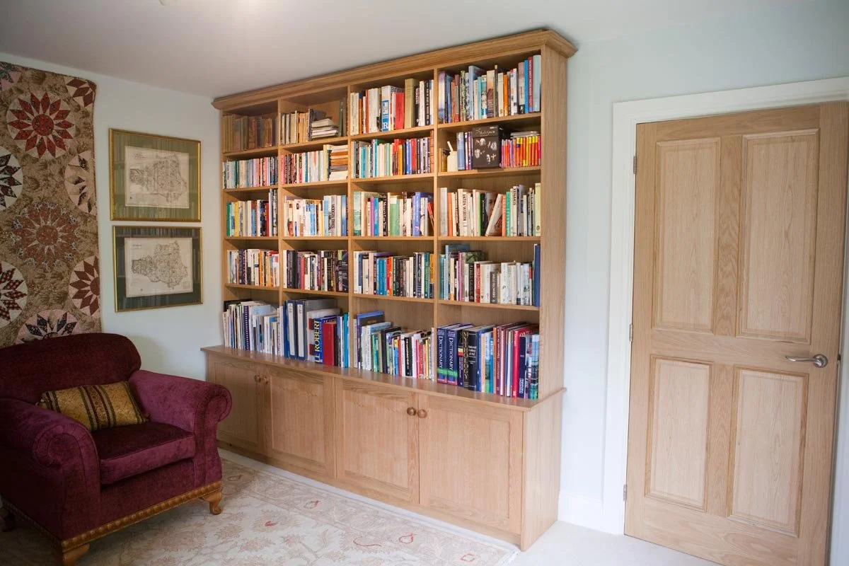 A wooden bookshelf filled with books in a living room, beside a red upholstered armchair and framed pictures on the wall.