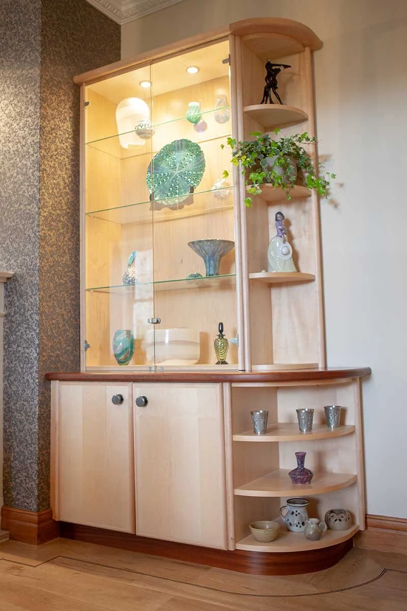 Wooden display cabinet with glass doors and multiple shelves showcasing decorative glassware, pottery, and figurines, and featuring additional shelving on the side with small sculptures and plants.