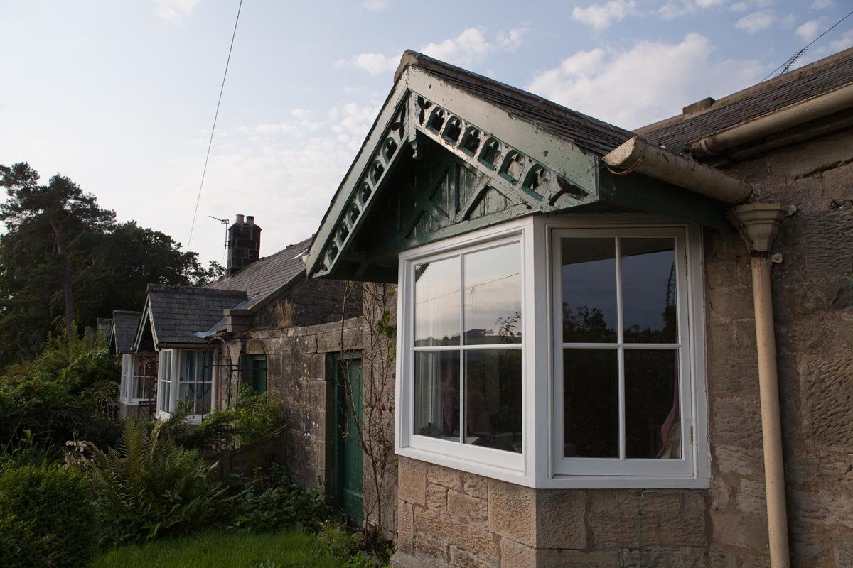 The exterior of a stone house with two bay windows, one with modern white frame and the other with older style. The house has a sloped roof and decorative wooden trim under the roof gable. The yard has bushes and grass, and the sky is partly cloudy.