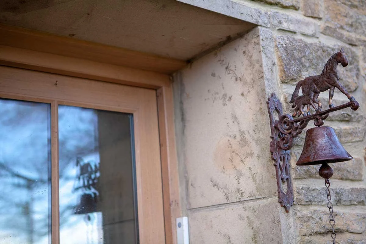 Rusty decorative bell with a horse and foal sculpture on top, mounted on a brick and stone wall next to a window.