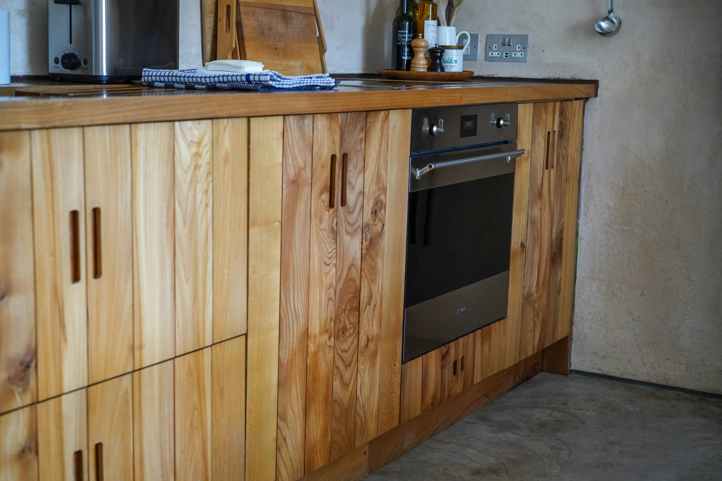 Wooden kitchen cabinets with a built-in oven, a countertop with a microwave, dish towel, and cooking essentials, and part of a wall with electrical outlets and a pipe.
