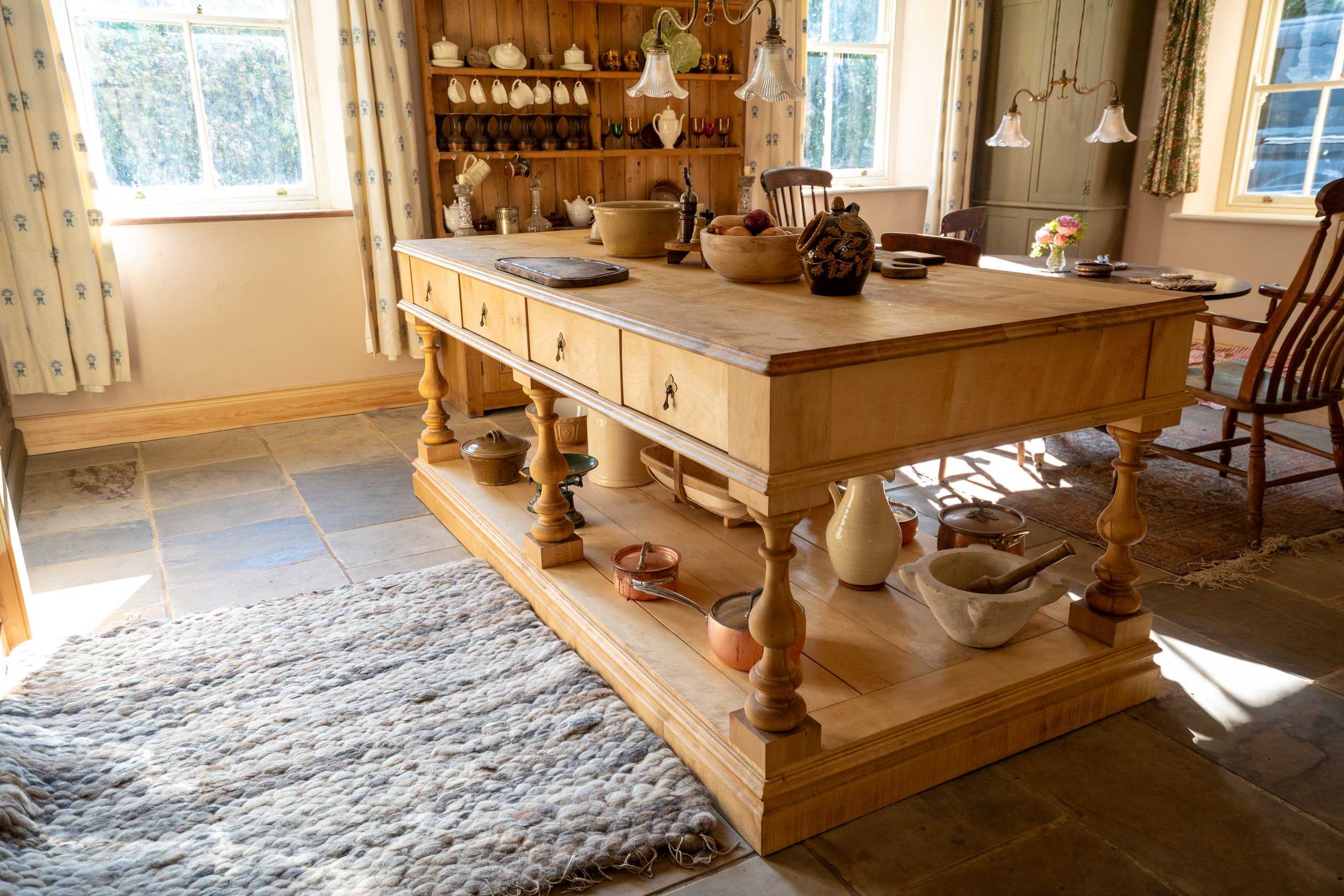 A rustic kitchen interior with a wooden island in the center, decorated with pottery and bowls, surrounded by natural light from windows, patterned curtains, and vintage-style furniture.