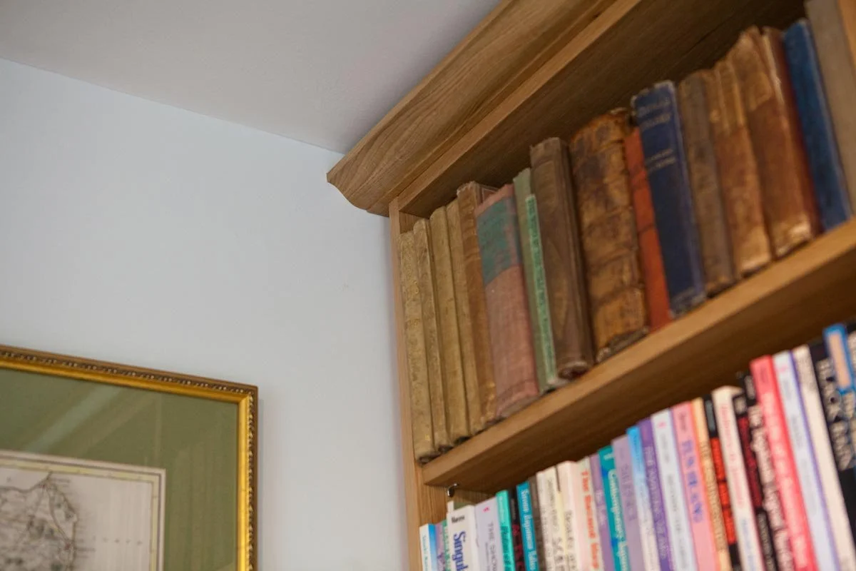 Close-up of a wooden bookshelf filled with old, worn books, with part of a framed map or artwork on the light-colored wall next to it.