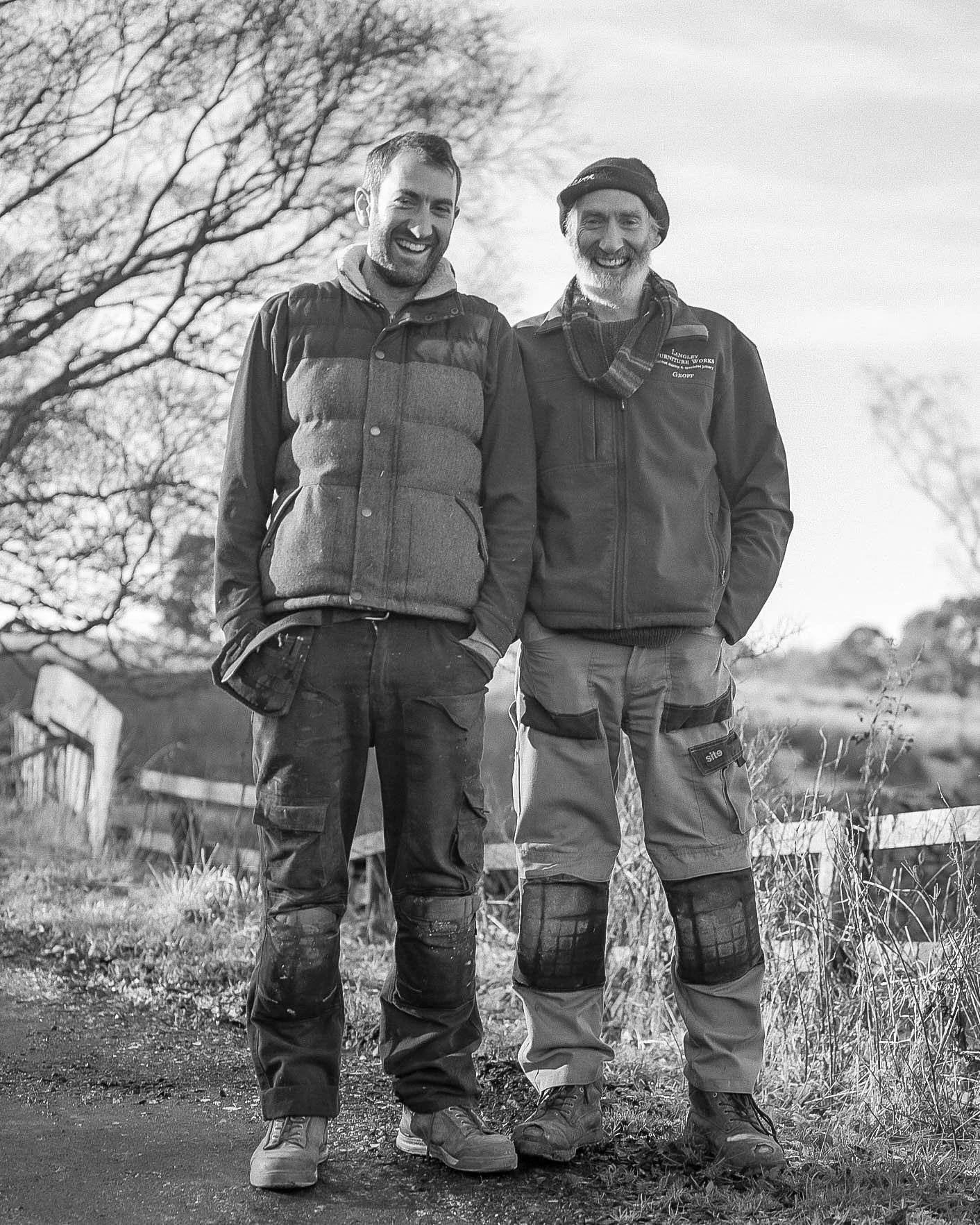 Two men standing outdoors on a dirt path, smiling, dressed in outdoor and work clothing with trees and a fence in the background.