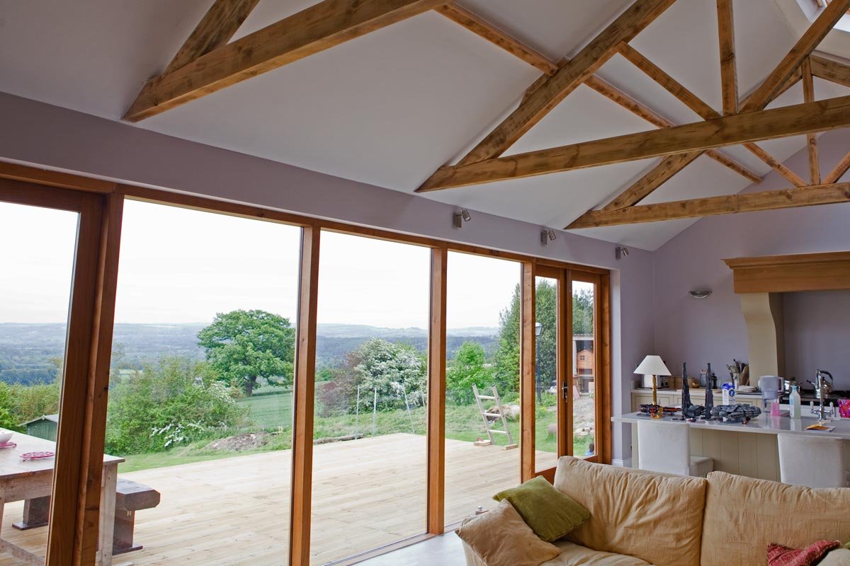 Living room with large floor-to-ceiling windows overlooking a green outdoor area with trees and hills, wooden ceiling beams, and a beige sofa with a green pillow.