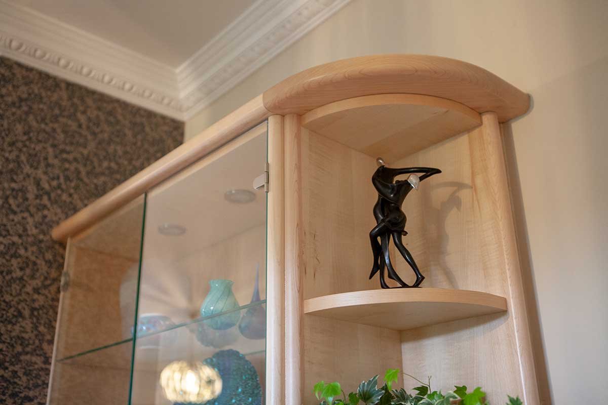 A wooden display cabinet with glass doors on the left and curved shelves on the right. The top shelf on the right contains a black cactus-shaped sculpture, and the lower shelf has a small green plant. The background shows a beige wall and a ceiling w