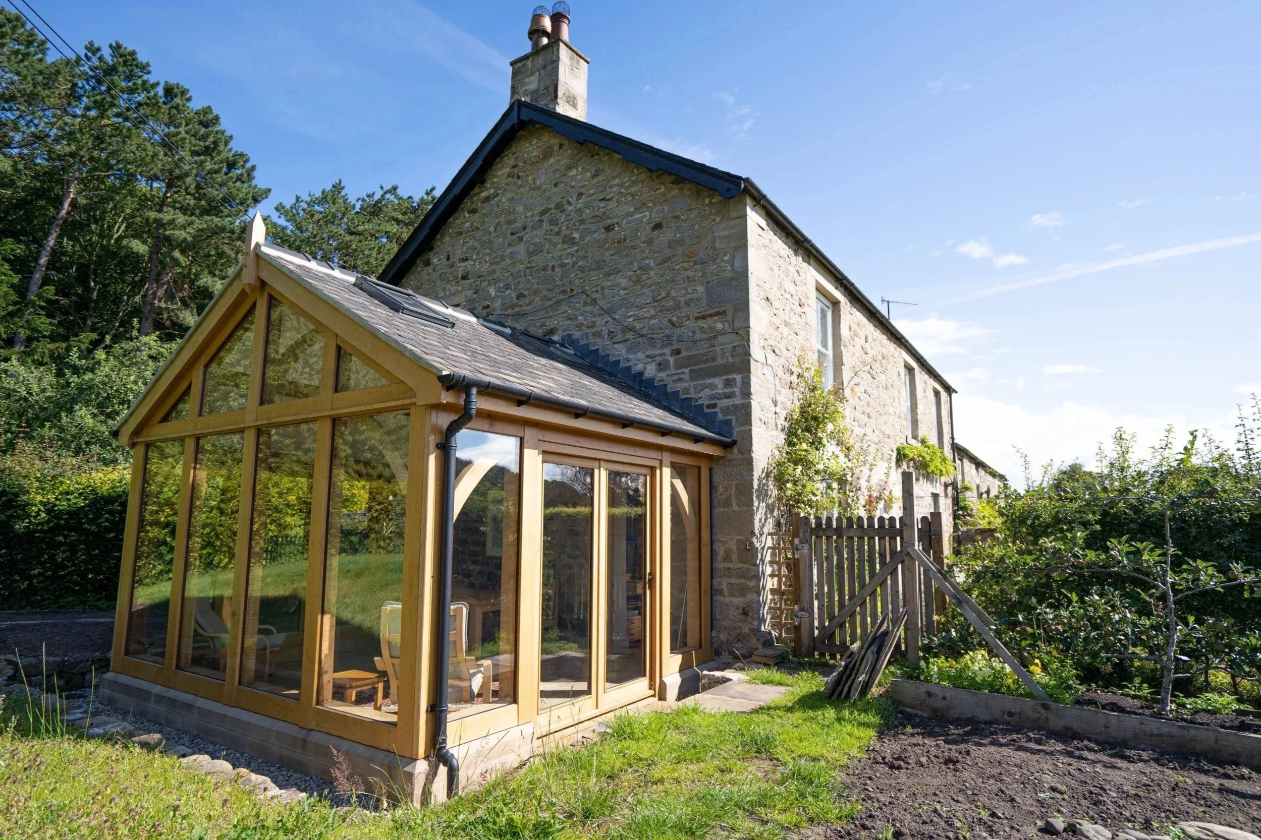 Stone house with a small wooden outbuilding and a garden, surrounded by trees, with a clear blue sky.