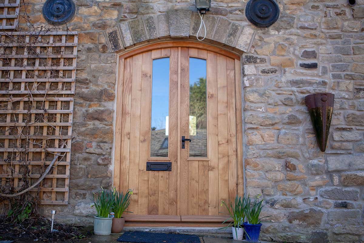 Wooden arched door with glass panels on a stone wall exterior, flanked by potted plants, with a wall-mounted light fixture and decorative wall sconces