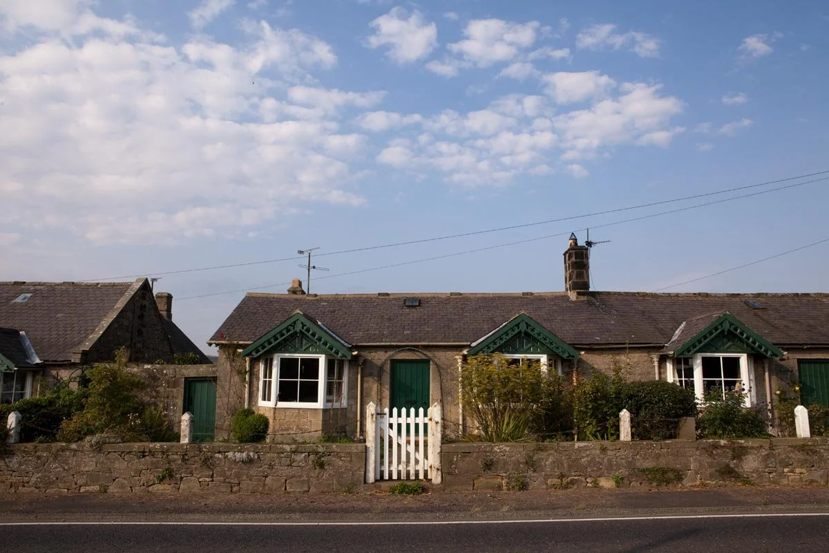 A row of old stone houses with green shutters and doors, small gardens in front, and a wooden gate, under a partly cloudy sky.