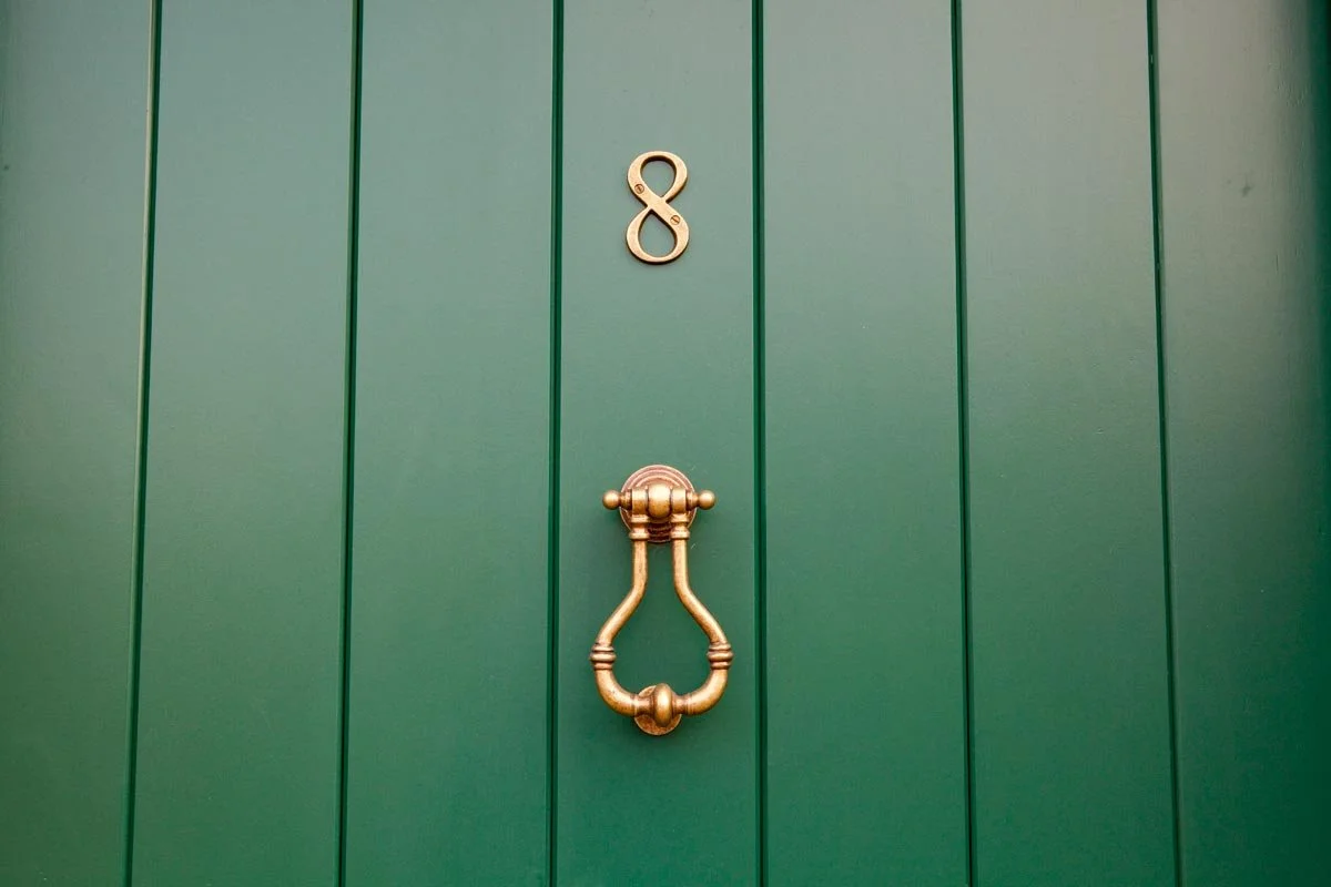 Green door with gold house number 8 and a gold door knocker shaped like a medieval ring.