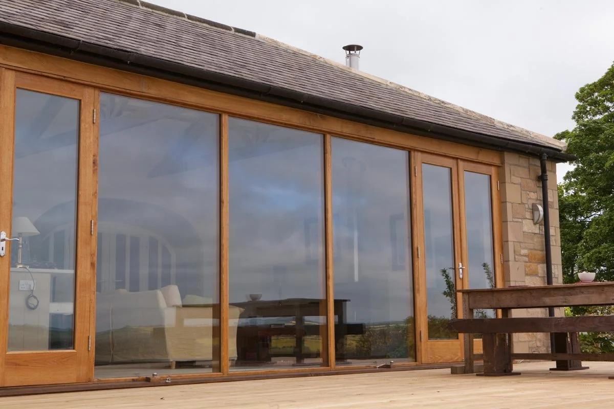 A house with large glass sliding doors framed in wood, a stone wall on the right, and a wooden deck outside with a bench and potted plants.