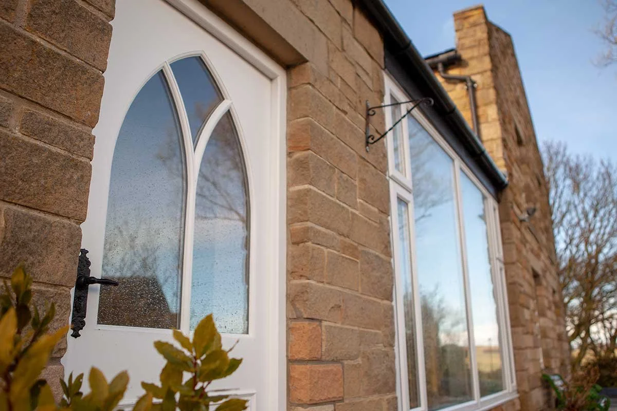 Close-up of a white front door with decorative glass panels next to a large window on a brick house. Christmas lights are hanging from the roof, and trees are visible in the background.