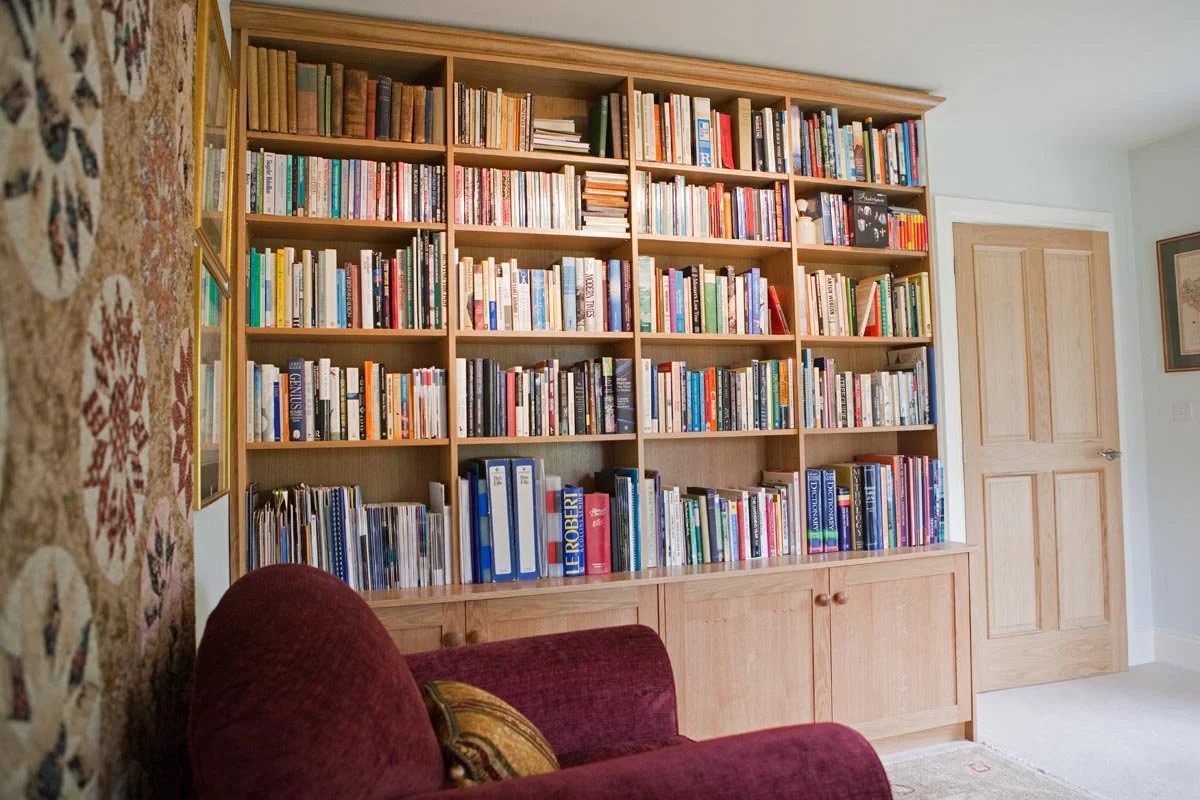 A large wooden bookshelf filled with various books in a living room. A maroon couch with pillows is partially visible in the foreground, and a light-colored door is on the right side of the image.