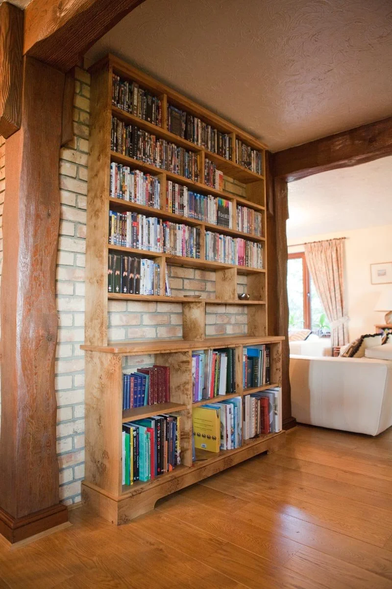 Wooden bookshelf filled with books in a cozy living room with wooden floors and brick wall, near a window with curtains.