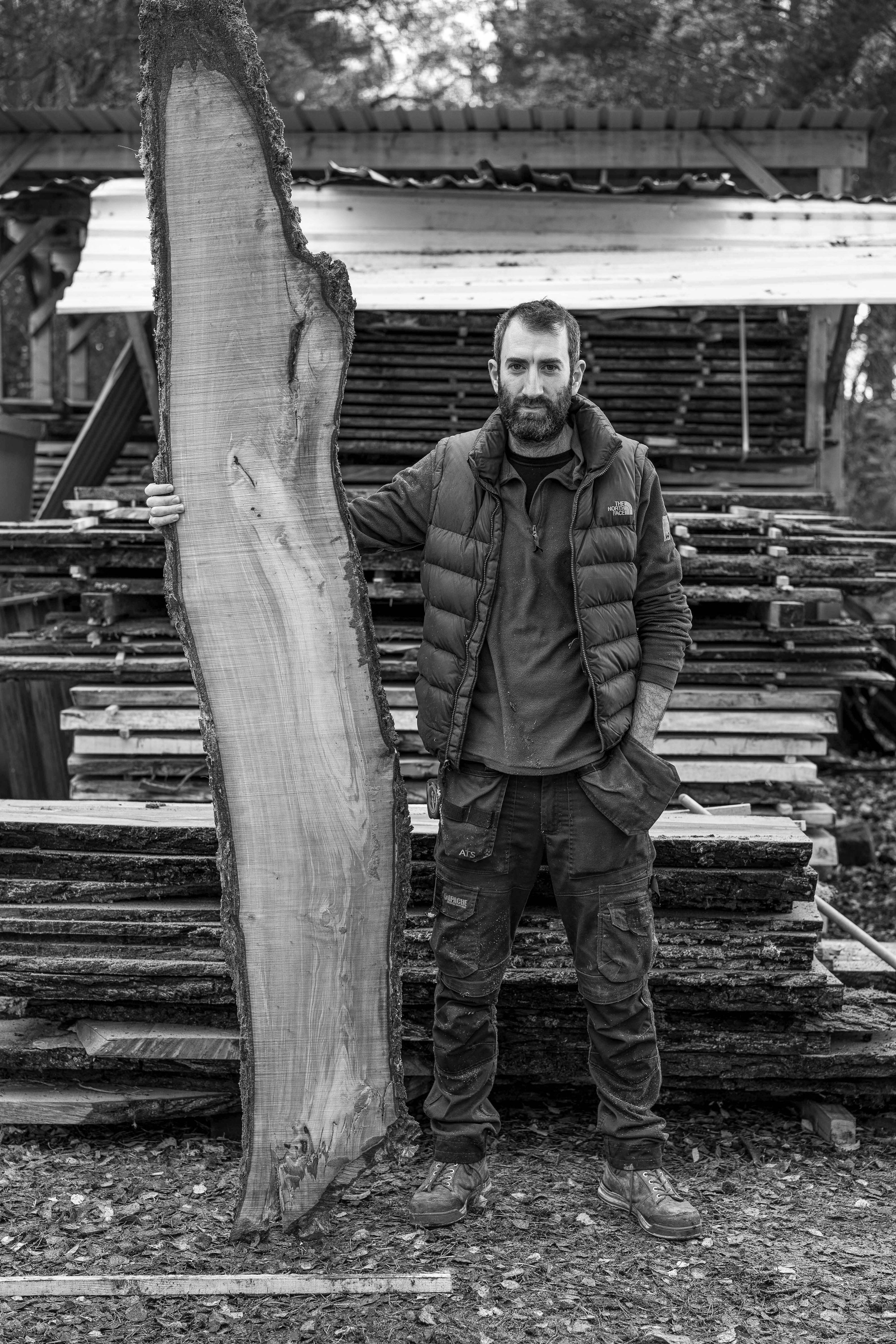 A man with a beard, wearing outdoor work clothes and a puffy vest, stands outdoors near a stack of cut wood. He leans against a large, freshly cut tree trunk.