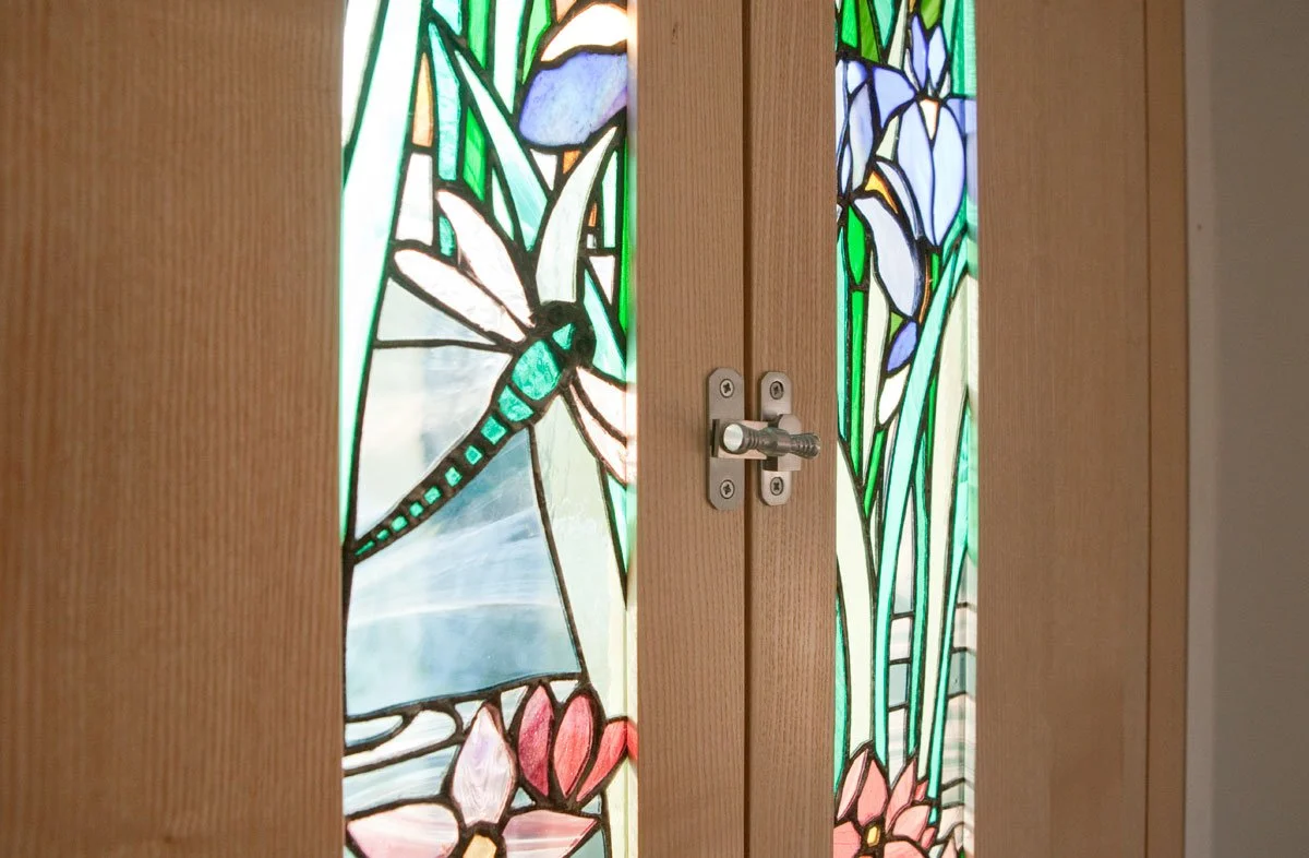 Stained glass door panels featuring a dragonfly, iris flowers, and other floral design, framed by wooden doors with a latch in the center.