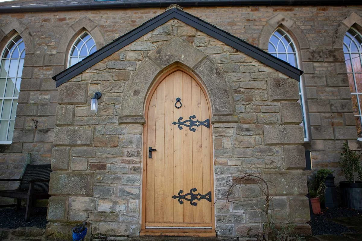 Wooden door with black iron hinges and handle, set in a stone wall with an arched opening, flanked by tall windows with pointed arches.