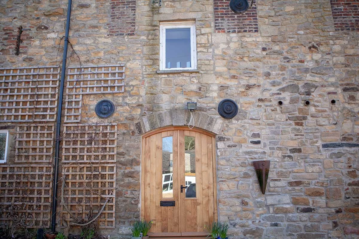 A rustic stone exterior wall of a building with a wooden arched door, a small window above, and garden plants in front. There are four black circular vents on the wall and a lattice on the left side.