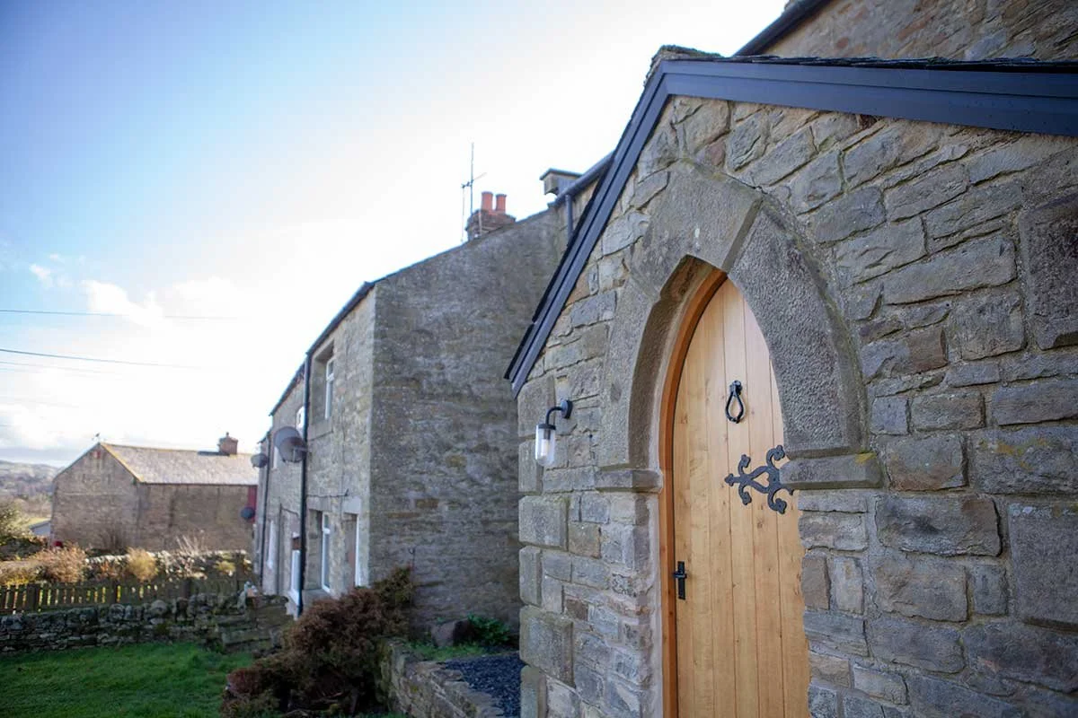 Stone cottage with wooden arched door, black iron hardware, and gray roof against a clear sky.