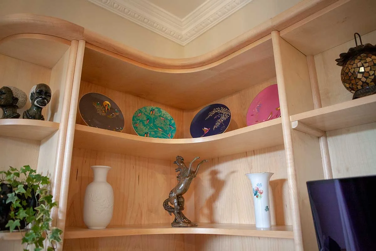 A wooden display shelf with decorative items including three black bust sculptures on the left, four decorative plates with colorful designs on the middle shelf, and two vases along with a bronze horse statue on the bottom shelf. A potted plant is pa