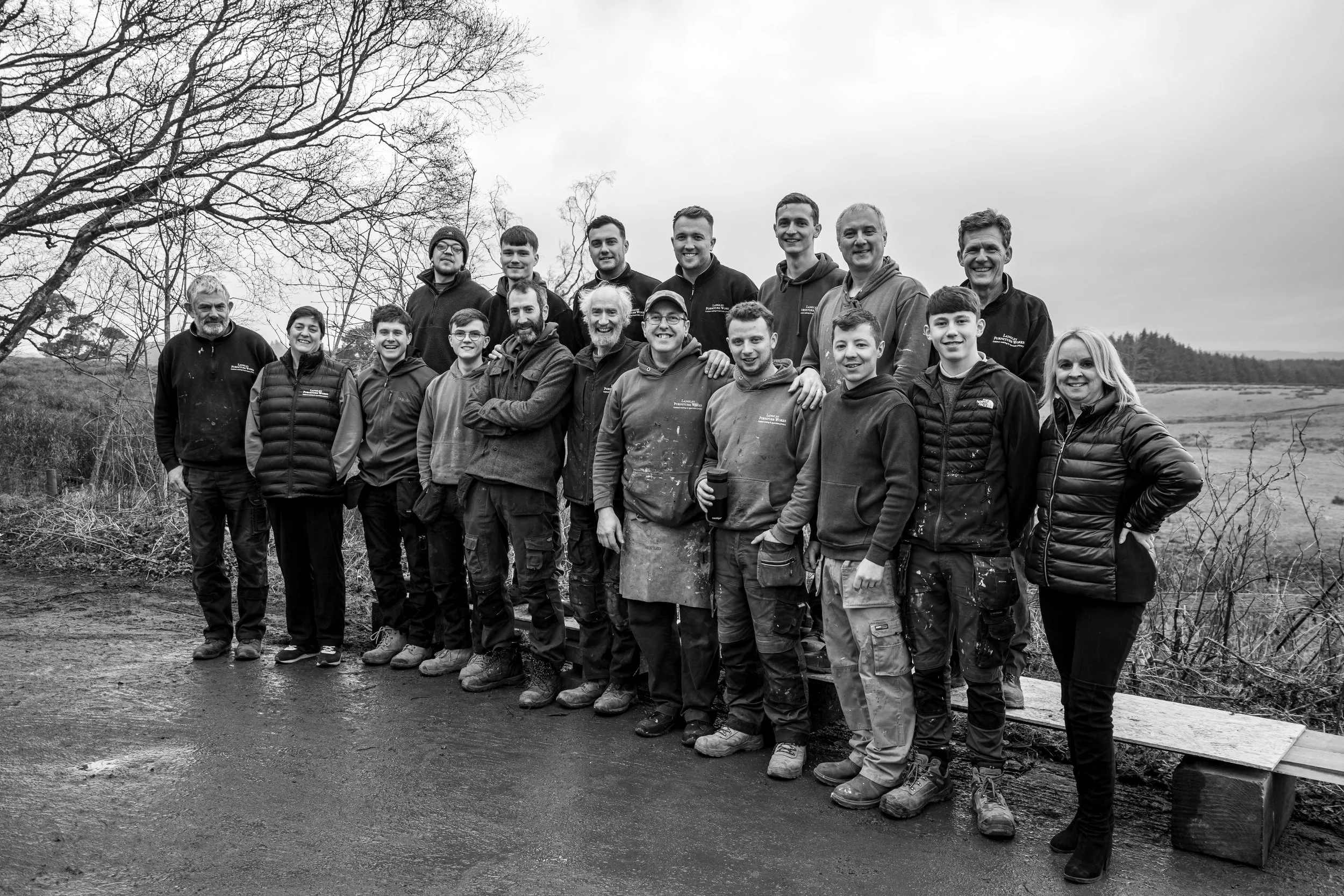 A group of 17 people standing outdoors on a rural path with fields and trees in the background of a cloudy day, some wearing work clothes and jackets.