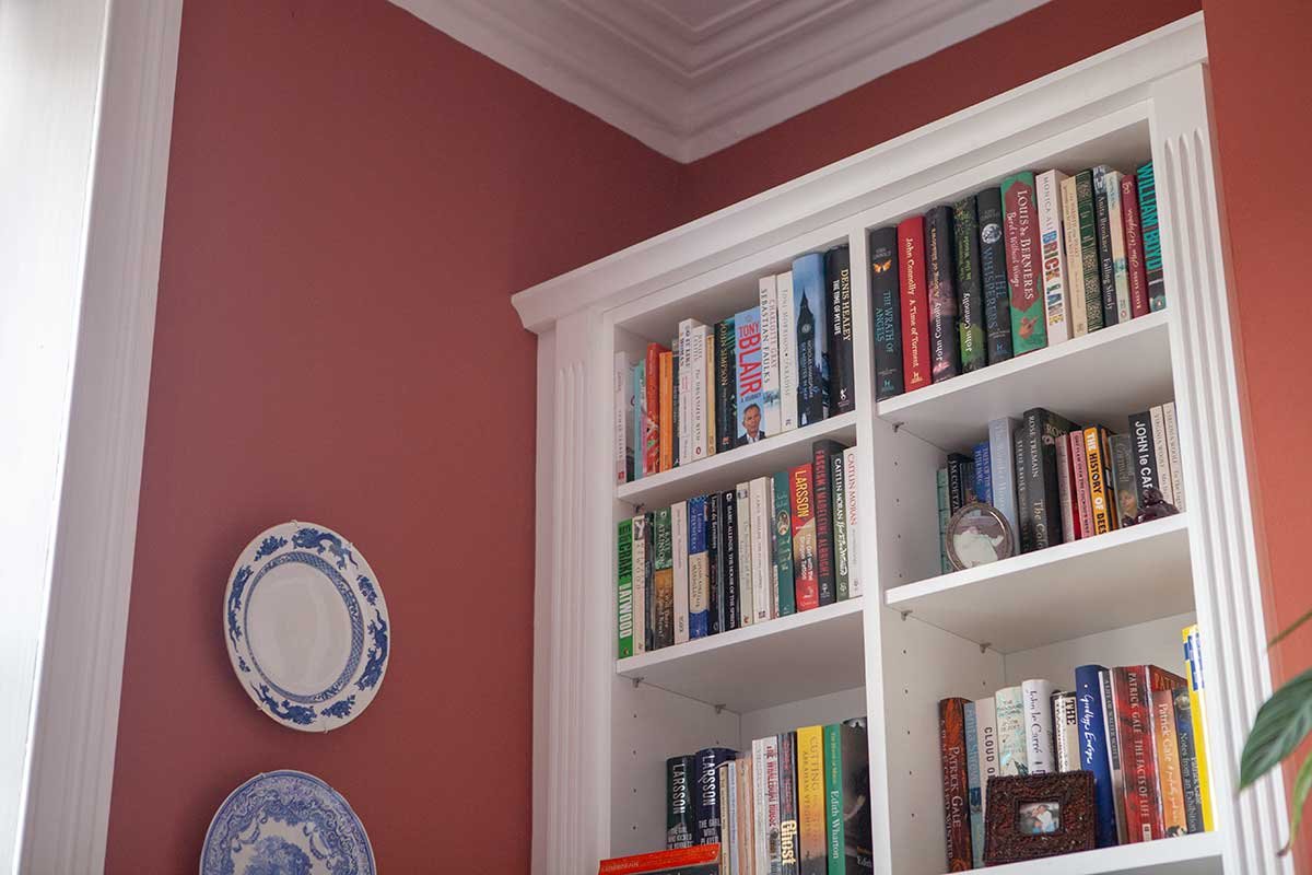 A white bookshelf filled with books against a pink wall with two decorative plates hanging on it.
