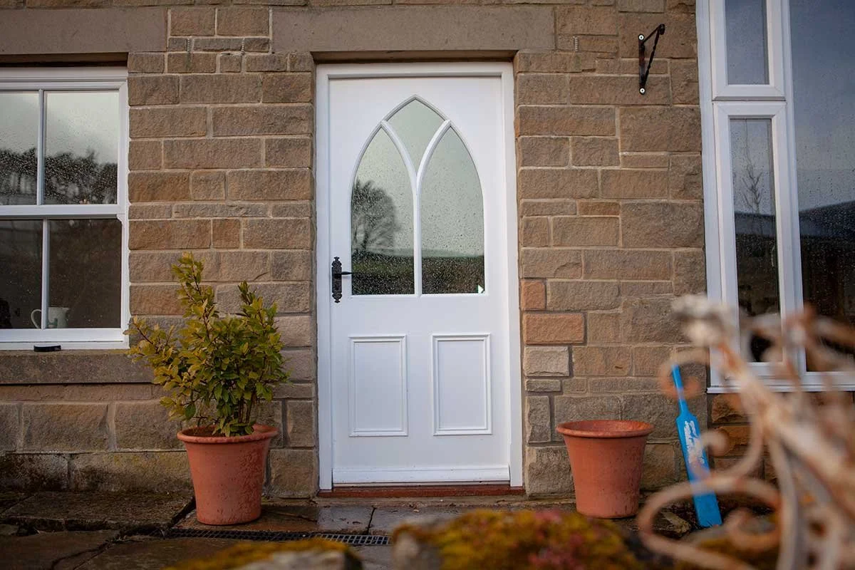 White front door with arched glass panels, flanked by two potted plants, situated on a brick wall exterior of a house.