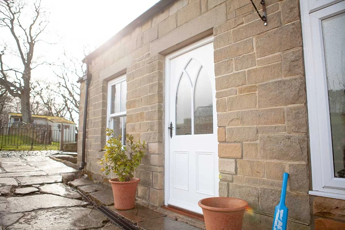 Front stone house with a white door, two potted plants, a blue shovel, and a stone pathway.