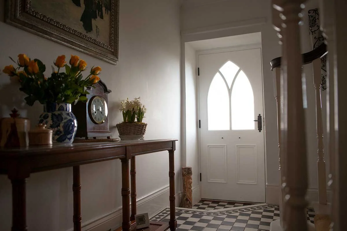 View of a narrow hallway with a white front door featuring a decorative glass window, a wooden console table with a vase of flowers, a small clock, and potted plants, underneath a framed painting. The floor has a black and white checkered tile patter
