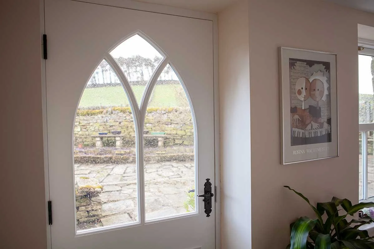 Interior view of a front door with two tall, narrow arched windows, showing a backyard with stone steps, grass, and stone walls, and a framed art print hanging on the wall beside the door.