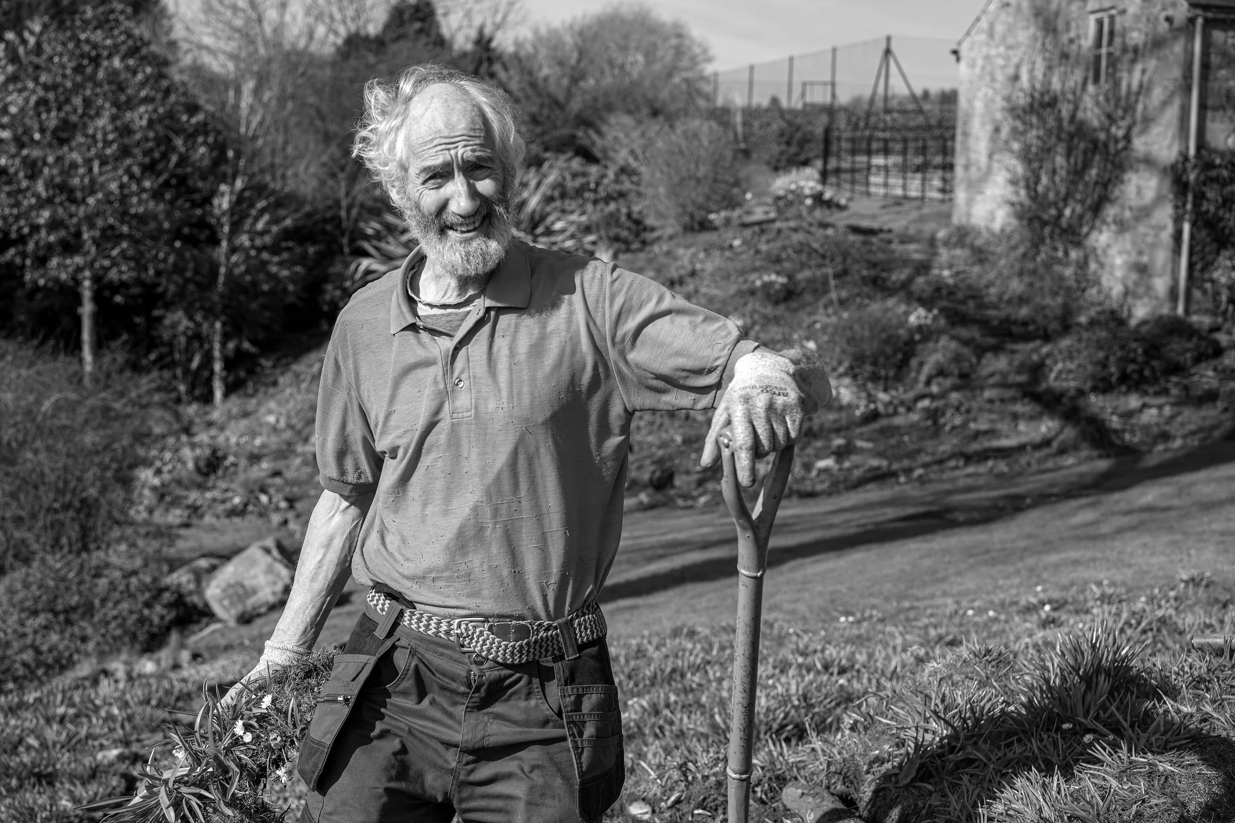 An elderly man with a beard and mustache smiling outdoors. He is wearing gardening gloves, a polo shirt, and a belt, and is holding a gardening tool. The background shows a garden or yard with plants, a building, and a fence.