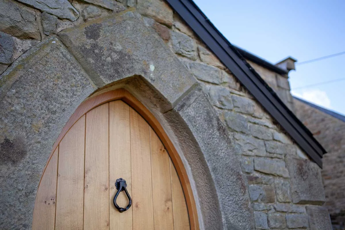 Close-up of a wooden door with a black metal knocker, set in a stone building wall with a sloped roof and blue sky in the background.