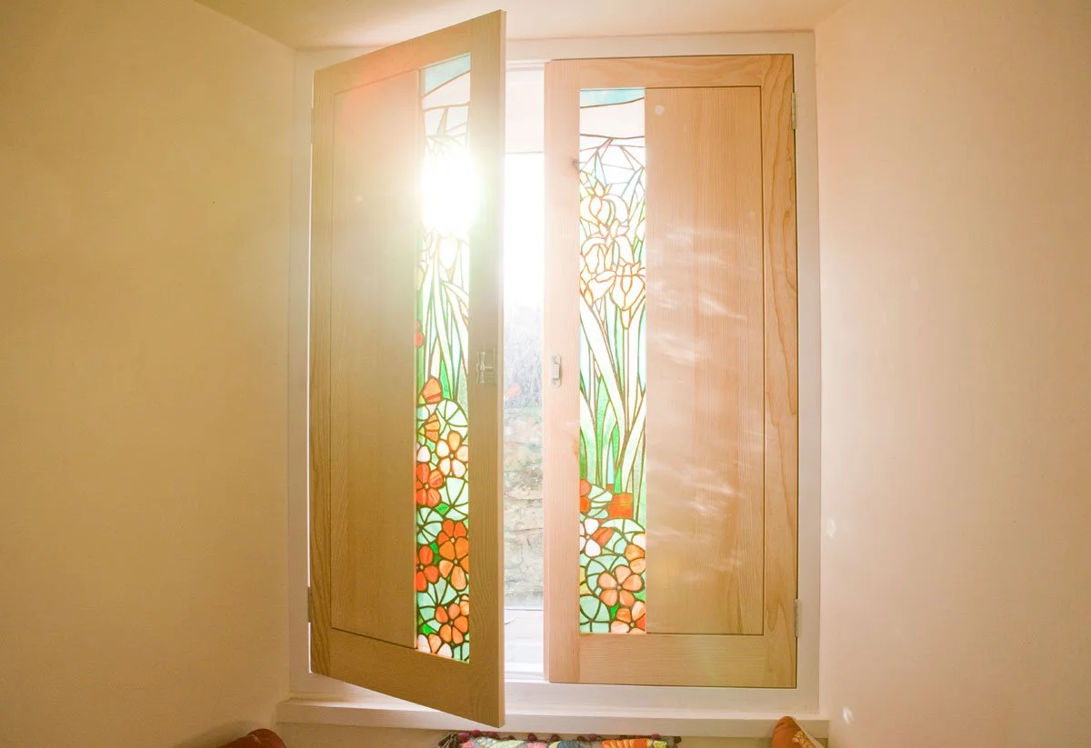 Open, wooden window with stained glass panels featuring floral and leaf designs, sunlight shining through