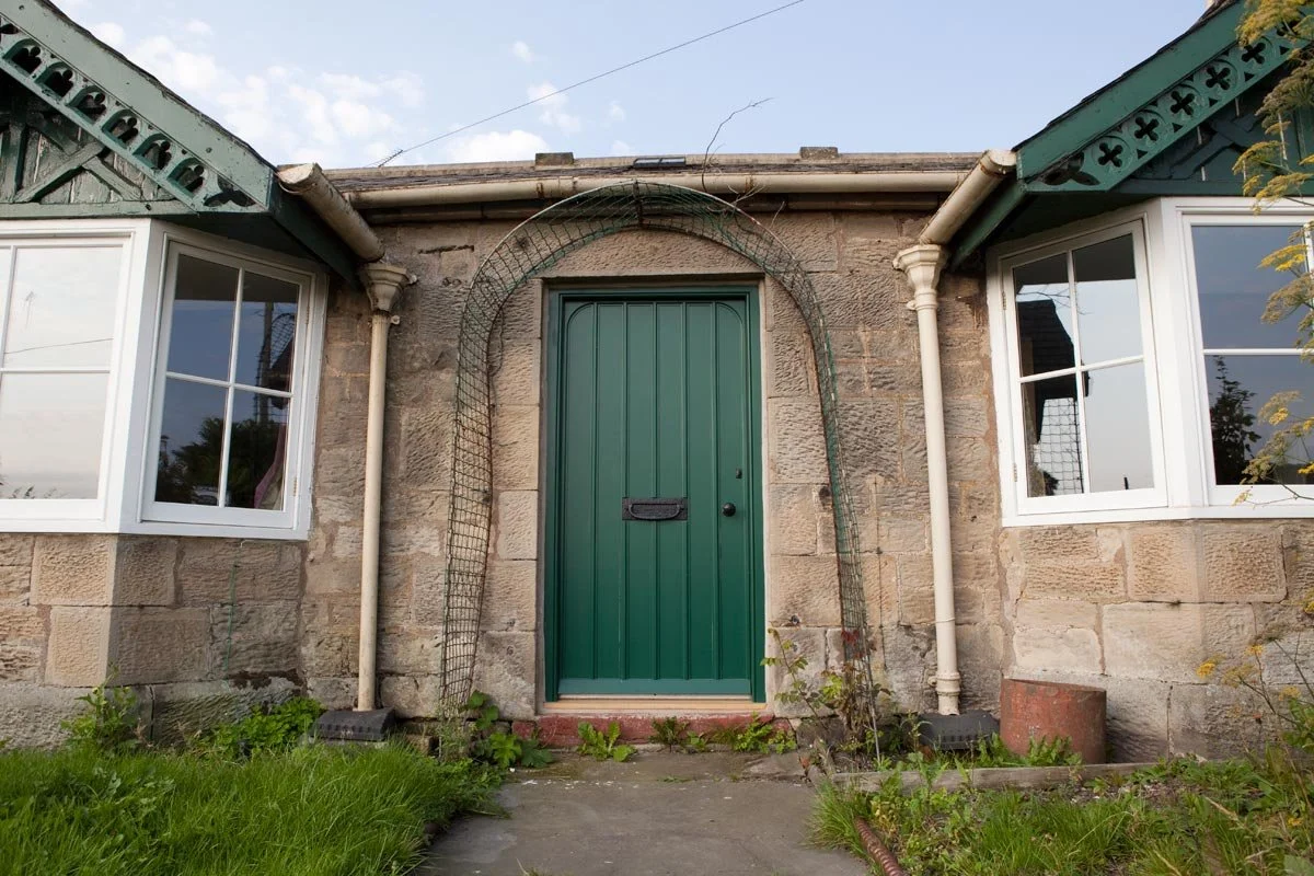 Front view of an old stone house with two large white-framed windows, a green door, and a small garden with grass and plants.