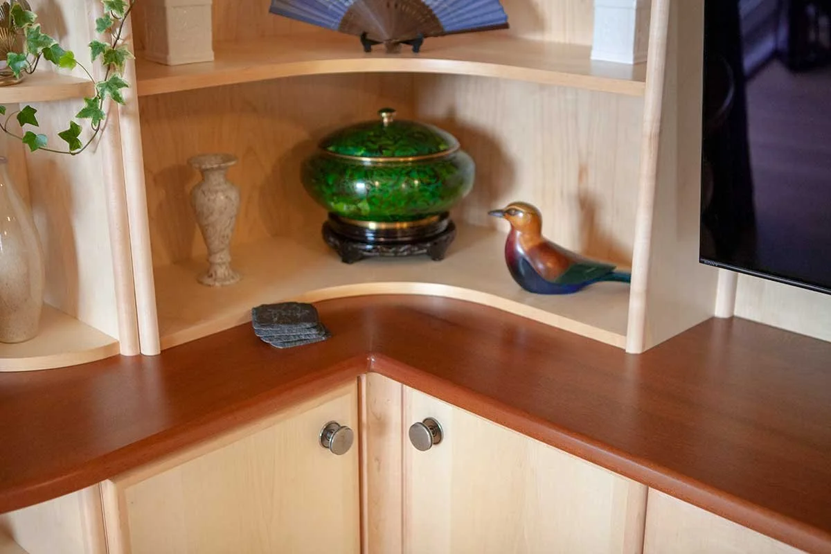 Wooden corner shelf with decorative items including a green bowl, a ceramic vase, a bird figurine, a small stack of black stones, and a partially visible potted plant with green leaves.