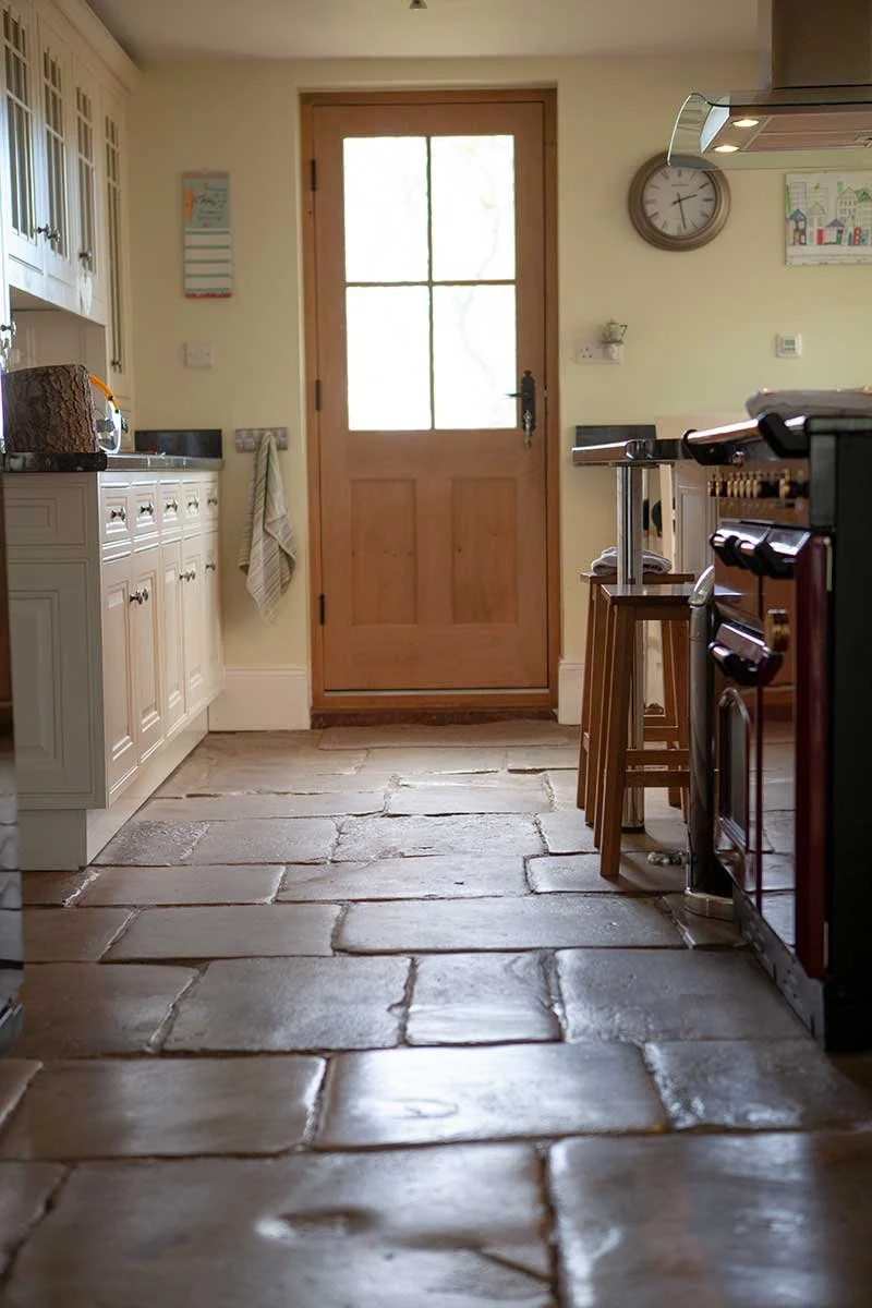 View of a kitchen entryway with a wooden door, stone tile floor, white cabinets, a wall clock, a small decorative object, and a counter with bar stools.