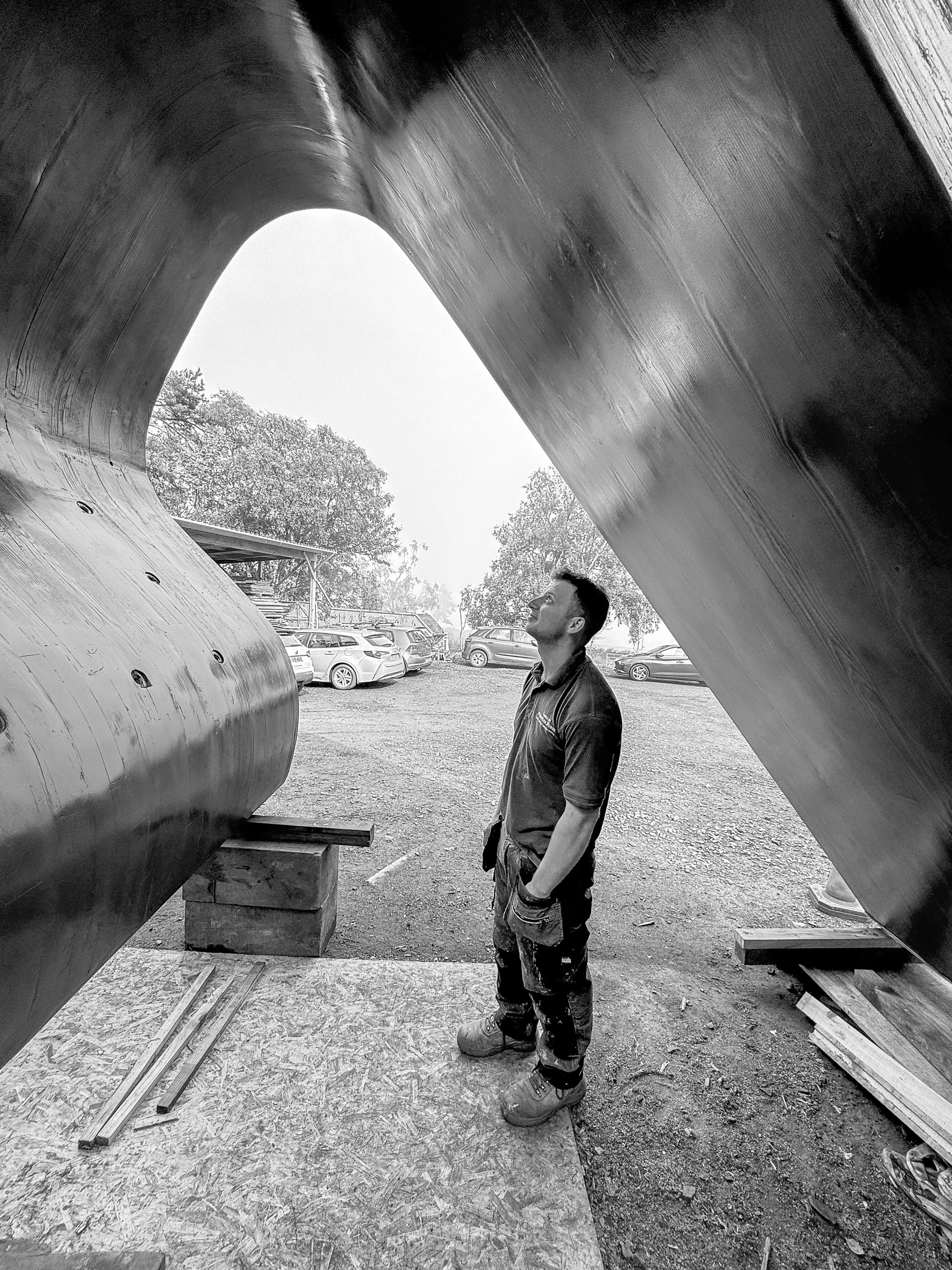 A man standing inside a large, curved, metallic sculpture or structure outdoors, with cars parked in the background.
