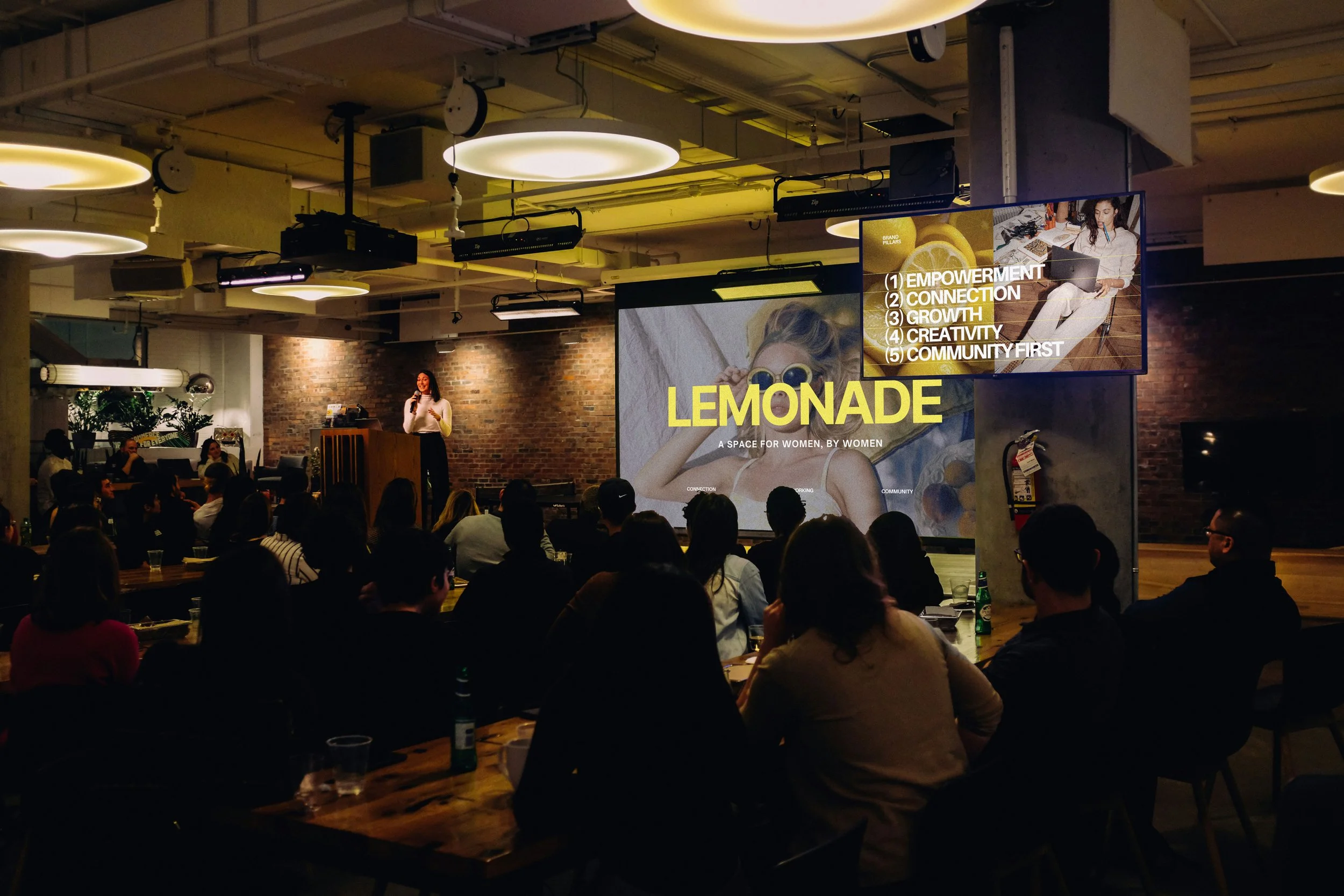 People attending a presentation or workshop in a large, modern indoor space with exposed brick walls and wooden tables. A woman stands at a podium addressing the audience, with a large screen displaying text about empowerment, connection, growth, creativity, and community, along with the word 'LEMONADE' and a subtitle about women by women.