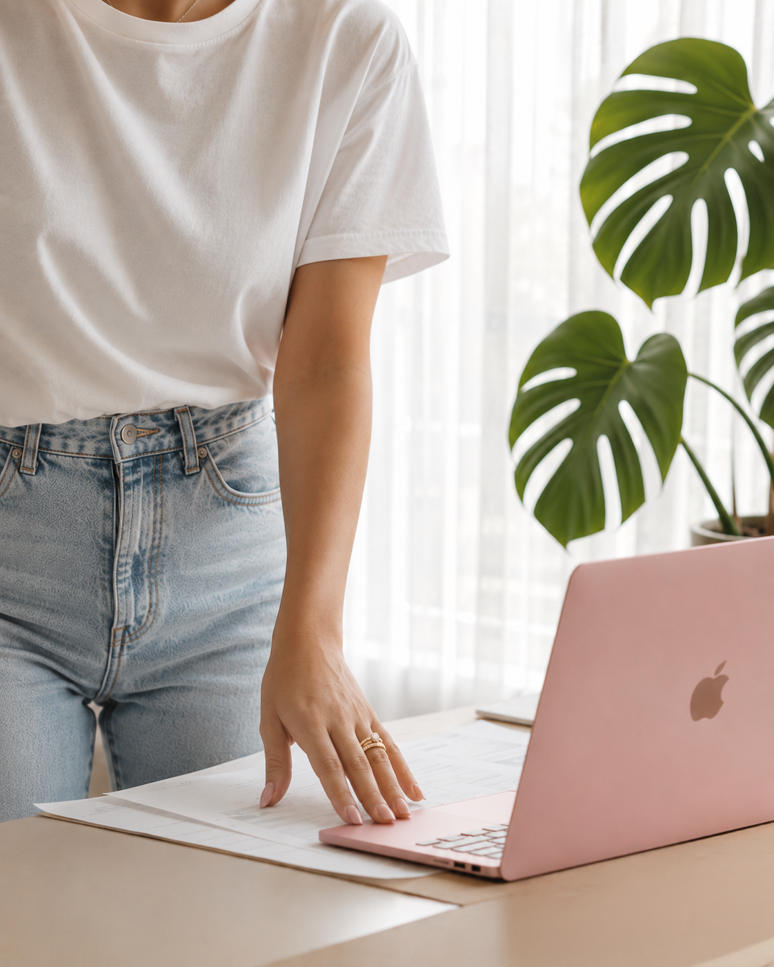 Person standing at a desk, touching papers, with a pink MacBook and a large green Monstera plant nearby.